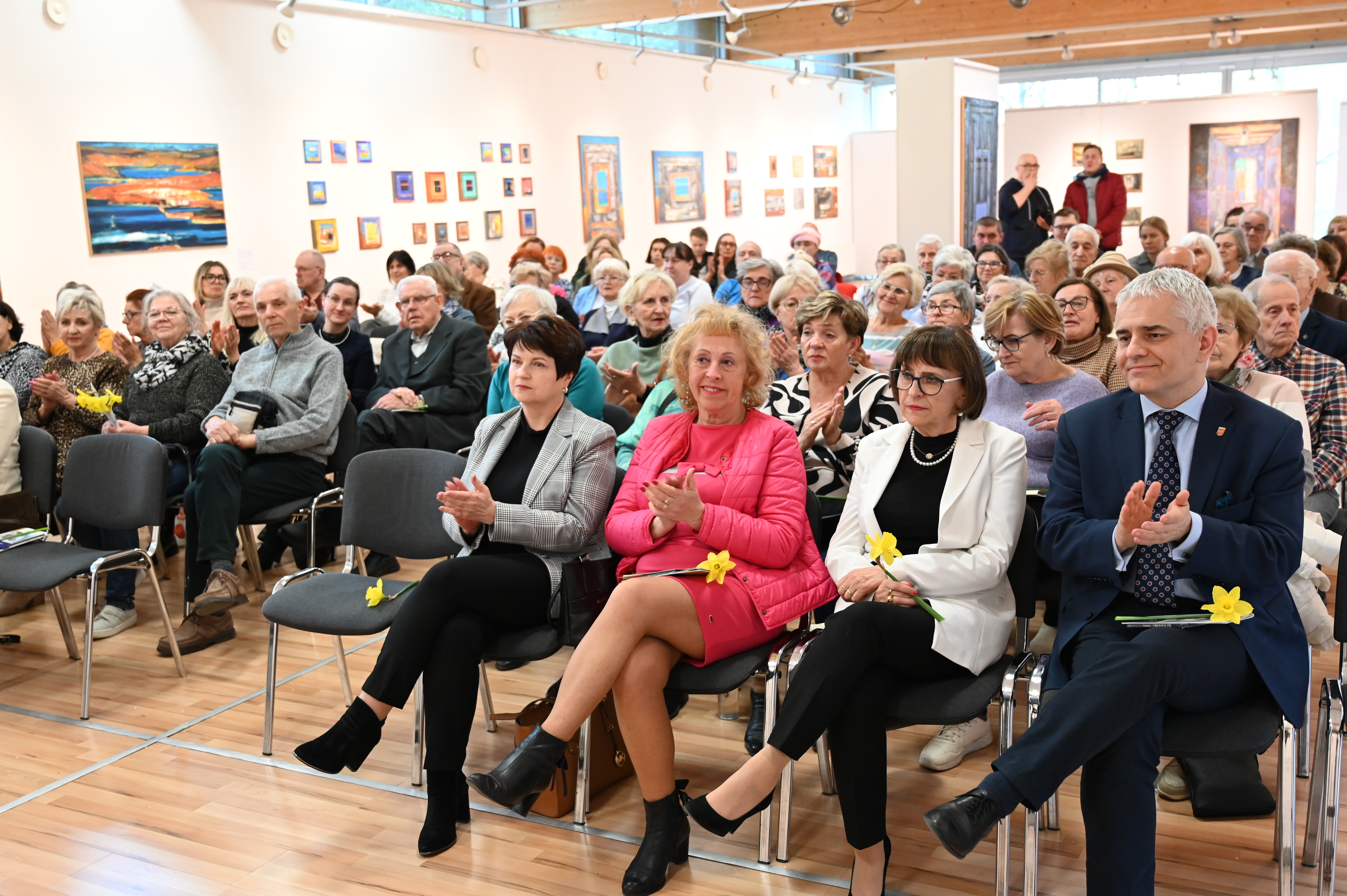 Pomieszczenie galerii z obrazami, w którym kilkadziesią dorosłych osób siedzi na krzesłach, słuchając prelekcji wygłaszanej przez kobietę. Mówczyni posługuje się wyświetlaną na ekranie prezentacją, w której zawarto informacjje o wsparciu społecznym dla osób dotkniętych chorobą Alzheimera i ich opiekunów.