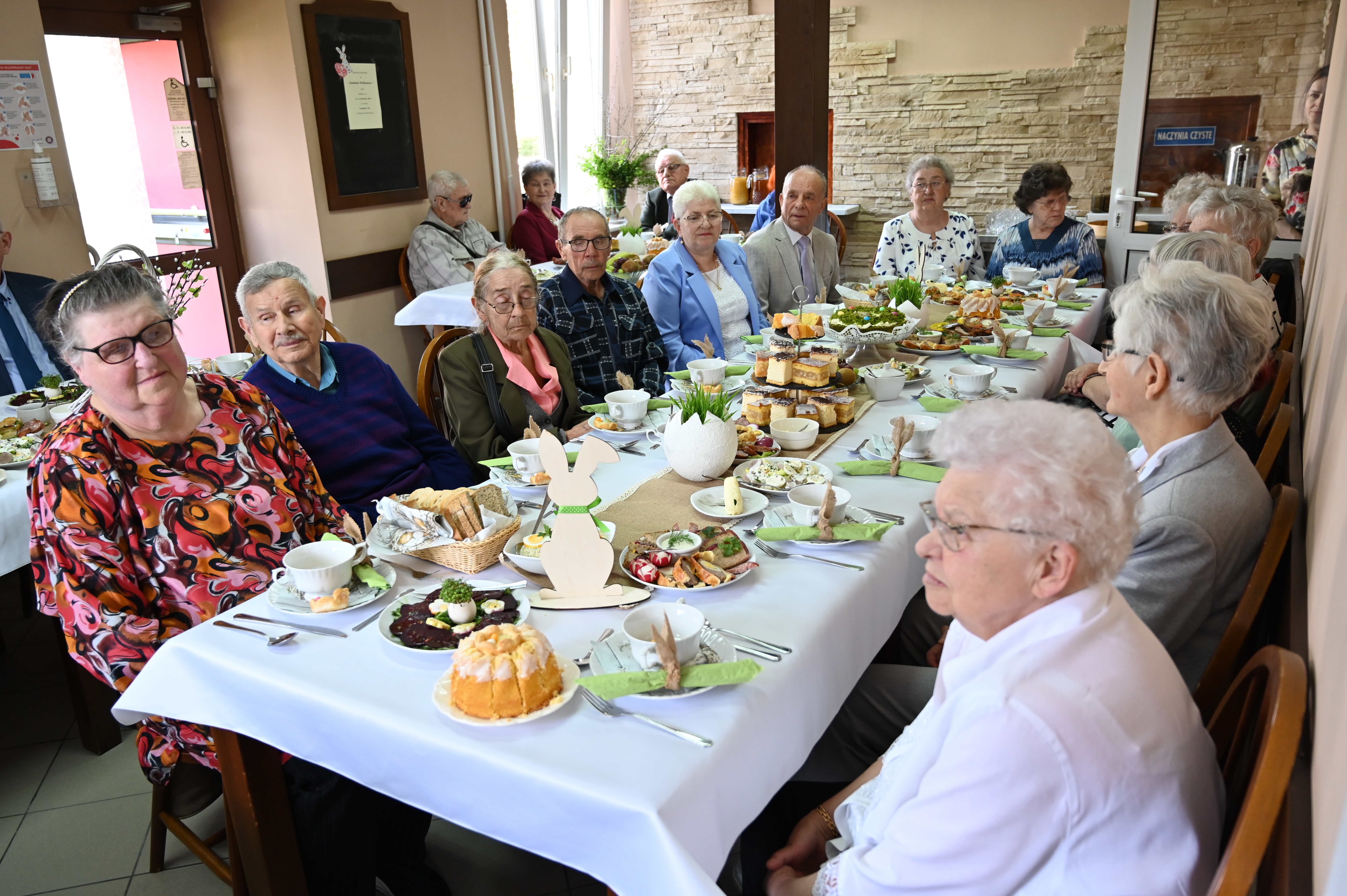 Zdjęcie przedstawia grupę senior&oacute;w: kobiet i&nbsp;mężczyn przy śniadaniu Wielkanocnym. Na stole potrawy wielkanocne.